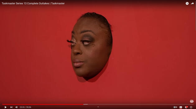 screenshot of a youtube video with the title and video controls visible. the titles reads: "Taskmaster Series 13 Complete Outtakes | Taskmaster." Judi Love is poking her head through a hole in a red panel with an unamused/disappointed expression.